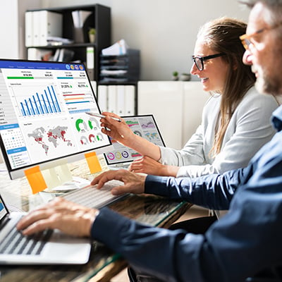 Photograph of two people working and collaborating, looking at a computer screen and discussing supply chain management.