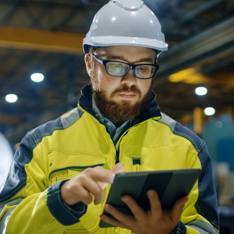 A man in a reflective jacket and hard hat reviews project information on a tablet