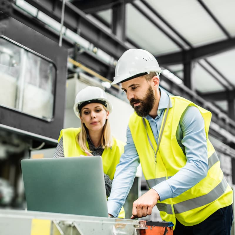 Two people in vests and hard hats review information on a computer in a factory setting
